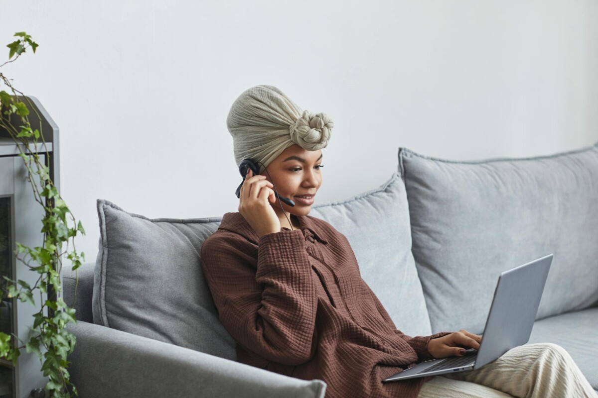 Woman wearing a headset and turban, working on a laptop from the couch.